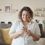 A person with curly hair is smiling while looking at their smartphone. They are sitting on a couch with decorative pillows in a cozy living room.