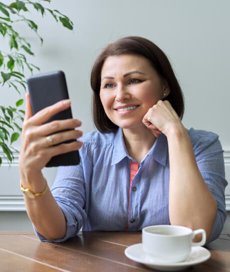 Woman smiles at smartphone while sitting at a table with a cup of coffee.