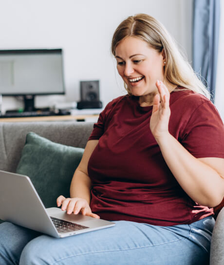 Woman in a maroon shirt sitting on a couch, smiling and waving at a laptop.