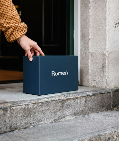 A hand picks up a dark blue Rumen box from a stone doorstep.