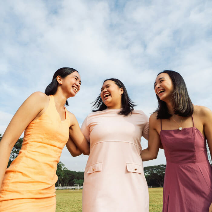 Three women in colorful dresses stand outside, smiling and embracing, with a grassy field and cloudy sky in the background.