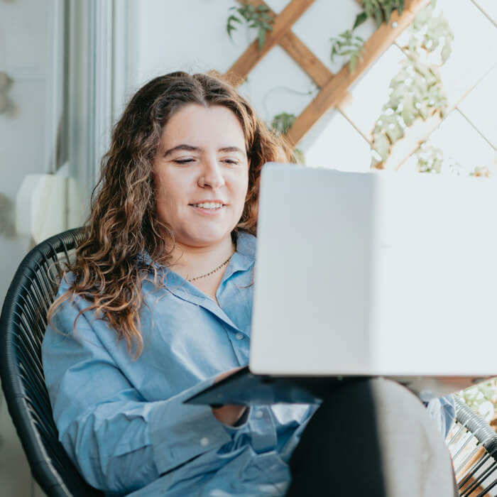A person with curly hair sits in a chair, using a laptop and smiling.