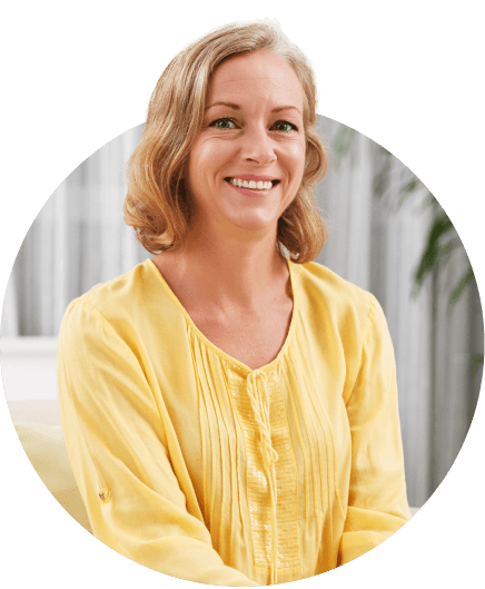 Smiling person with shoulder-length hair wearing a yellow blouse, seated indoors against a blurred background.