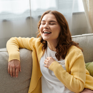 A person with long hair wearing a yellow cardigan and white shirt sits on a sofa, laughing with one hand on their chest.