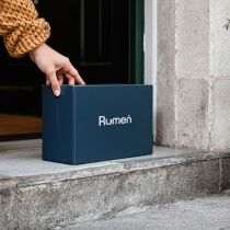 A person wearing a textured sweater picks up a dark blue box labeled "Rumen" from a doorstep.